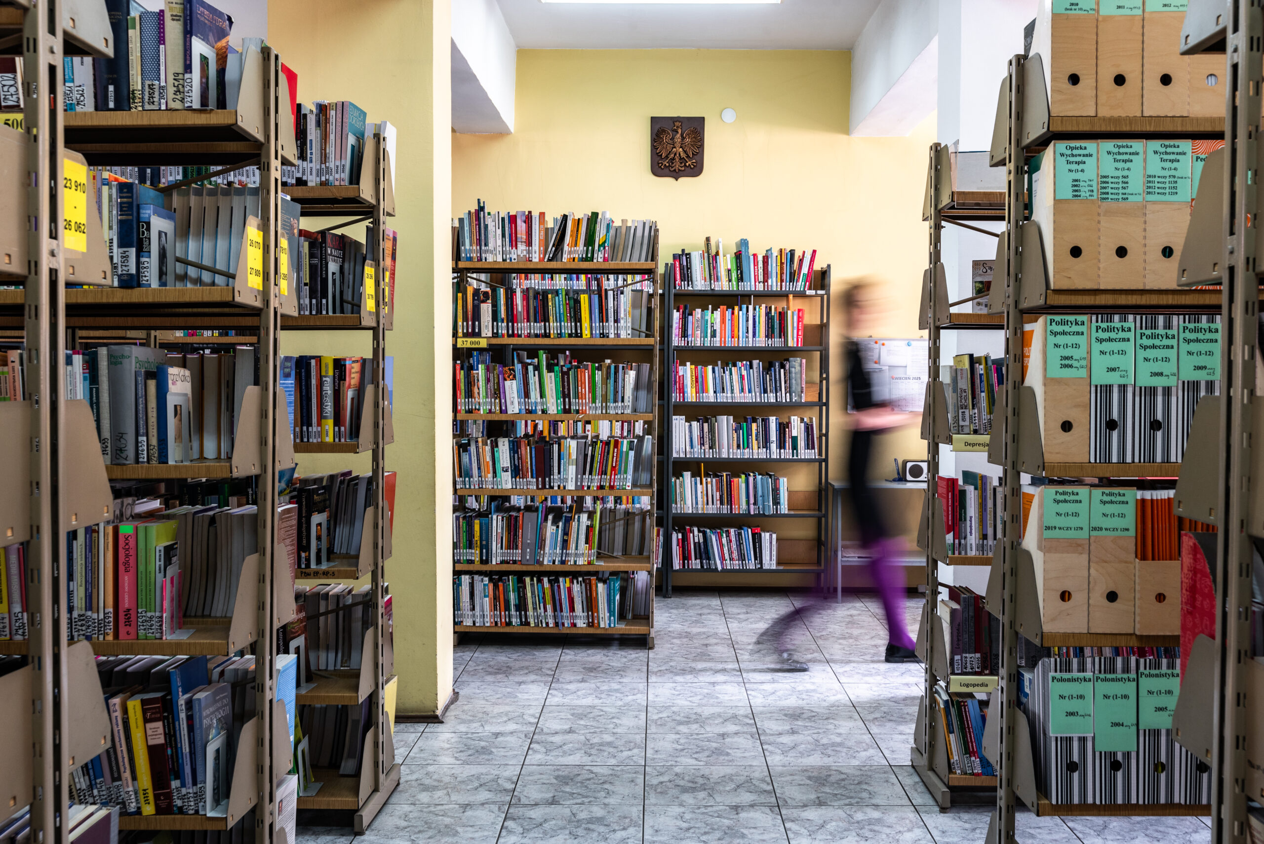 Wnętrze magazynu bibliotecznego w Żyrardowie z rzędami metalowych regałów. Regały są szczelnie wypełnione książkami oraz segregatorami z czasopismami (m.in. „Polityka Społeczna”). Na żółtej ścianie w głębi wisi godło Polski. Podłoga wyłożona jest jasnymi płytkami. Na zdjęciu uchwycono rozmazaną sylwetkę osoby w ruchu, co nadaje kompozycji dynamiki